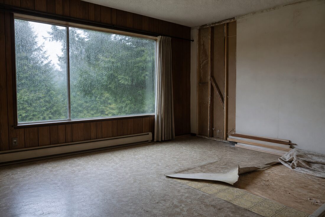 Living room of a 1970s Fraser Valley home part way through a renovation, with sheet vinyl flooring peeled back to reveal an older layer of vinyl tile, a textured stipple ceiling above and rain running down the window